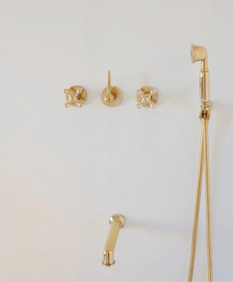 Pode incluir: Torneiras douradas para casa de banho contra uma parede branca. O conjunto inclui um chuveiro com mangueira flex&iacute;vel, um bico curvo e tr&ecirc;s comandos de parede. Os acess&oacute;rios t&ecirc;m um design cl&aacute;ssico e elegante.