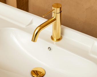 May include: A polished gold-tone bathroom faucet with a single lever handle mounted on a white ceramic sink basin. The sink features a matching gold-tone circular drain cover and a small white overflow drain hole located below the faucet spout.
