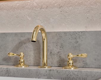May include: A polished gold bathroom faucet set featuring a high-arched spout and two separate lever handles. The fixture has a reflective metallic finish and is mounted on a gray stone surface. The text BRASSLIK is visible in the image.