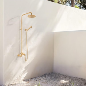 May include: A gold-colored outdoor shower fixture with a round showerhead and a handheld showerhead. The shower is mounted on a white wall, with a gravel floor and a glimpse of greenery.
