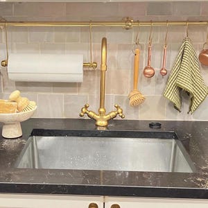May include: A kitchen sink area with a brass faucet, paper towel holder, and hanging utensils. A small basket with flowers and a soap dish are on the countertop. The background features a tiled backsplash and a brass rod with hanging items.