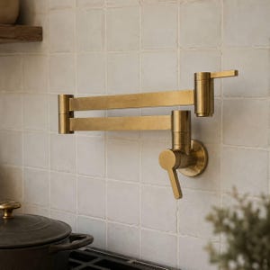 Könnte beinhalten: Ein wandmontierter Topffüller-Wasserhahn mit gebürstetem Messing-Finish, ausgestattet mit einem zweigliedrigen Schwenkarm und zwei Hebelgriffen zur Wassersteuerung. Die Armatur ist an einer hellen, quadratisch gefliesten Wand über einem Herd montiert.