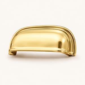 May include: A polished gold-toned metal cup pull handle featuring a smooth, rounded, semi-circular design with a reflective surface. The hardware has a curved, ergonomic shape with a recessed grip area and flat mounting ends on each side.