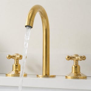 May include: A gold-colored bathroom faucet with a curved spout and two cross-shaped handles. Water flows from the spout, creating a visual of functionality and design. The faucet is set against a white background.