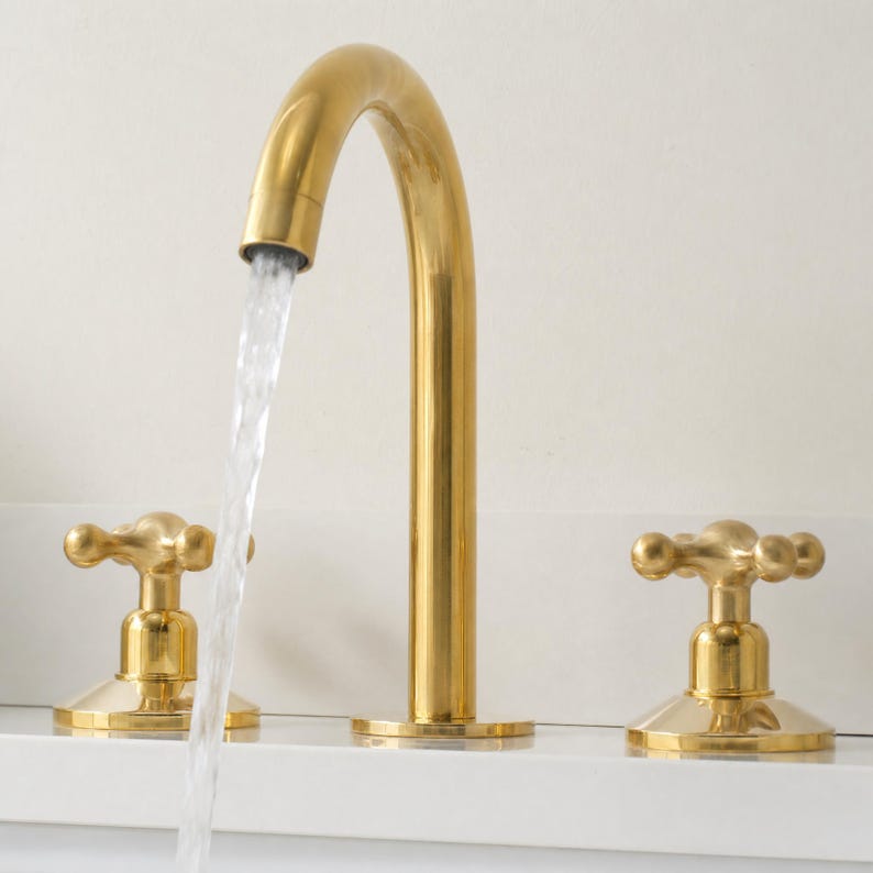 May include: A gold-colored bathroom faucet with a curved spout and two cross-shaped handles. Water flows from the spout, creating a visual of functionality and design. The faucet is set against a white background.