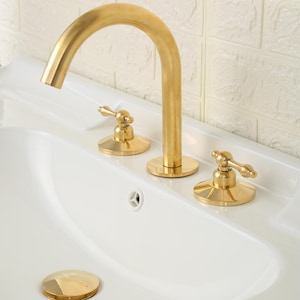 May include: A gold-toned bathroom faucet set featuring a high-arched spout and two separate lever handles mounted on a white ceramic sink basin. The set includes a matching gold-toned circular drain cover positioned at the bottom of the sink bowl.