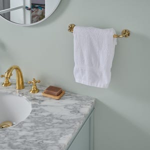 Puede incluir: Una escena de baño con una toalla blanca colgada de un toallero dorado. El lavabo tiene un grifo y accesorios dorados, y una pastilla de jabón descansa sobre una bandeja de madera. La encimera es de mármol y la pared es de color verde claro.