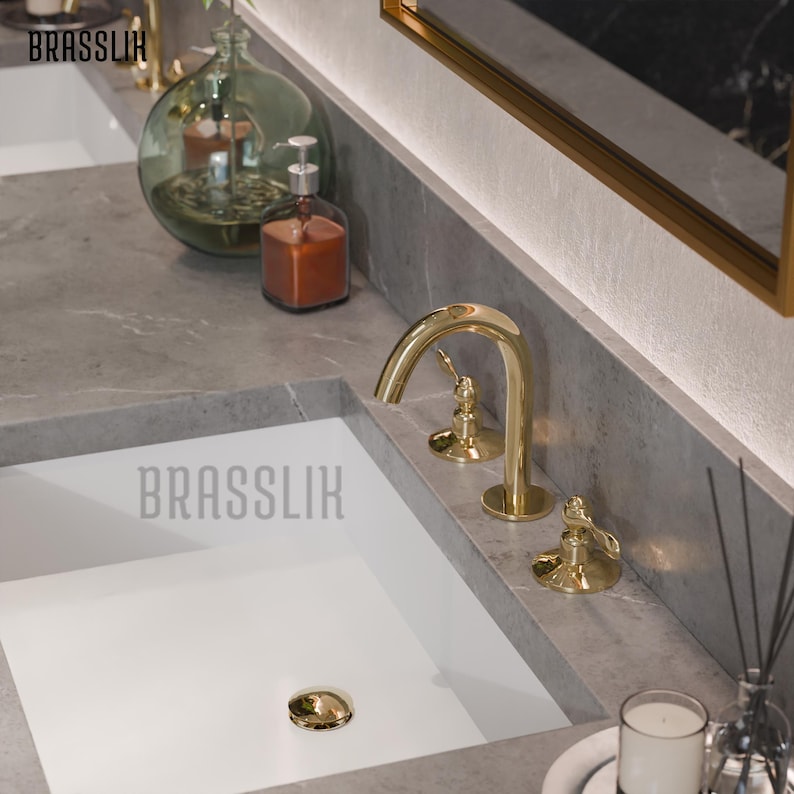 May include: A gold-colored bathroom faucet with a curved spout and two handles. The faucet is installed on a gray countertop above a white sink. The word "BRASSLIK" is printed on the sink.