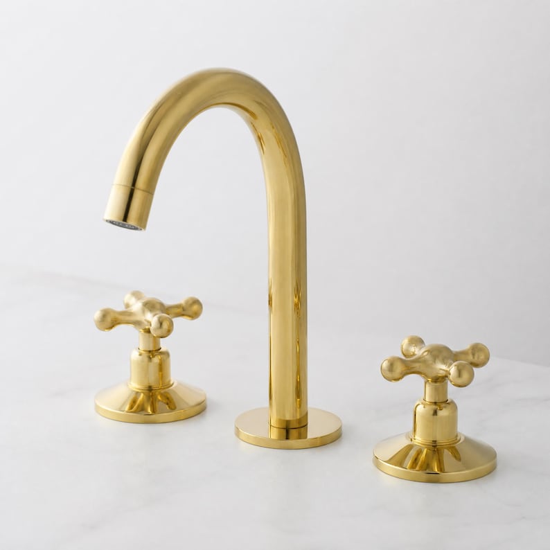 May include: A gold-colored bathroom faucet with a curved spout and two cross-shaped handles. The faucet is made of metal and has a shiny finish. The design is modern and elegant.