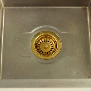 May include: A stainless steel kitchen sink with a gold-colored drain. The drain has a circular design with multiple slots for water drainage. The sink is clean and appears to be new.