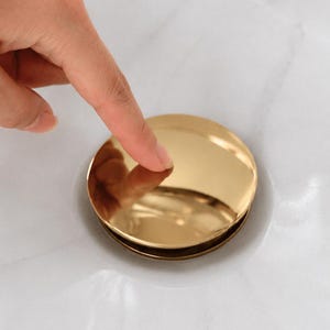 May include: A circular, polished gold-tone metal sink drain stopper with a smooth, reflective surface. The stopper is positioned within a white marble sink basin, featuring a simple, minimalist design that sits flush against the drain opening.
