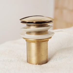 May include: A gold-toned bathroom sink drain stopper with a white plastic ring around the top. The stopper is made of metal and has a threaded design.