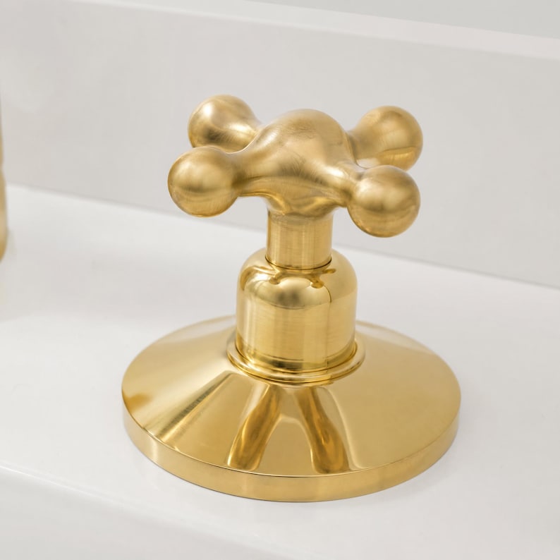 May include: A close-up of a gold-colored faucet handle. The handle has a cross-shaped design with four rounded knobs. The base of the faucet is circular and also gold-colored, reflecting the light. The faucet is set against a white background.