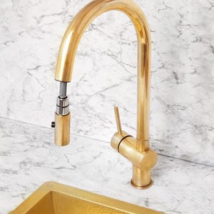 May include: A gold kitchen tap with a high, curved spout and a pull-down spray head. The tap features a single lever and is made of a shiny, metallic material. The background is a marble-effect surface.