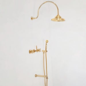 Pode incluir: Um conjunto de acess&oacute;rios de duche dourados contra uma parede branca. O conjunto inclui um chuveiro curvo, um chuveiro de m&atilde;o com mangueira e comandos de torneira. Os acess&oacute;rios t&ecirc;m um design cl&aacute;ssico e elegante.