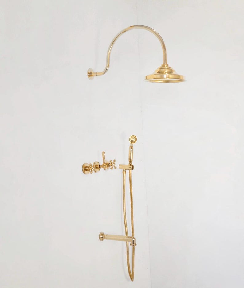 Pode incluir: Um conjunto de acess&oacute;rios de duche dourados contra uma parede branca. O conjunto inclui um chuveiro curvo, um chuveiro de m&atilde;o com mangueira e comandos de torneira. Os acess&oacute;rios t&ecirc;m um design cl&aacute;ssico e elegante.