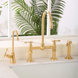 May include: A brushed gold kitchen faucet set featuring a high-arc bridge faucet with two lever handles, a separate smaller filtered water tap, and a matching side sprayer, all mounted on a white countertop above a white sink basin.