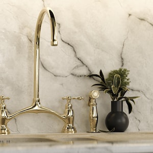 May include: A gold kitchen tap with two handles and a separate sprayer. The tap is mounted on a white marble worktop.