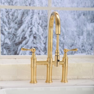 May include: A gold-colored kitchen faucet with a high-arc spout and two lever handles. The faucet has a pull-down sprayer and is set against a window with a snowy outdoor scene.