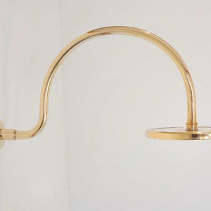 Pode incluir: Um bra&ccedil;o de chuveiro dourado com um design curvo. O bra&ccedil;o &eacute; montado em uma parede branca e possui um chuveiro circular. A lumin&aacute;ria &eacute; feita de metal e tem um acabamento polido.