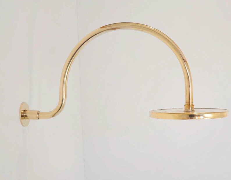 Pode incluir: Um bra&ccedil;o de chuveiro dourado com um design curvo. O bra&ccedil;o &eacute; montado em uma parede branca e possui um chuveiro circular. A lumin&aacute;ria &eacute; feita de metal e tem um acabamento polido.