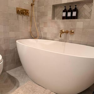 May include: A modern bathroom with a white oval bathtub, gold fixtures, and a built-in shelf with dark bottles. The walls and floor are tiled in neutral tones, and a beige bath mat sits on the floor.