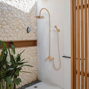 May include: A gold-colored shower fixture with a rain shower head and handheld showerhead. The shower is mounted on a white wall next to a stone wall and a wooden slatted partition. The showerhead is round.