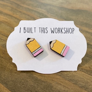 Puede incluir: Un par de pendientes de madera con forma de lápiz, pintados de amarillo y rosa. Los pendientes están sobre una tarjeta blanca con el texto "I Built This Workshop".
