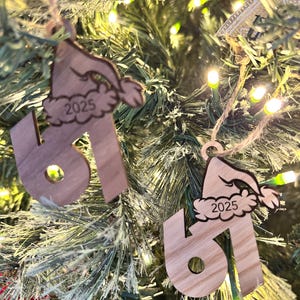 May include: Two wooden Christmas ornaments shaped like the number 61, each topped with a Santa hat and the year 2025. The ornaments are hanging on a Christmas tree with green needles and small, glowing lights.