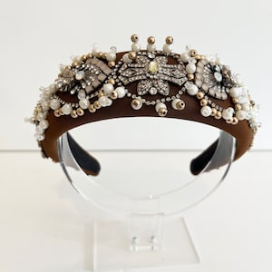 May include: A brown headband with a decorative design of gold and silver beads, rhinestones, and pearls. The headband is on a clear stand.