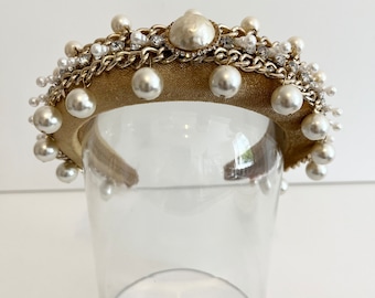 May include: A gold headband with a large pearl and chain detail. The headband is decorated with smaller pearls and rhinestones.