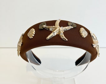 Puede incluir: Una diadema de terciopelo marrón con adornos de estrellas de mar y conchas doradas y plateadas.