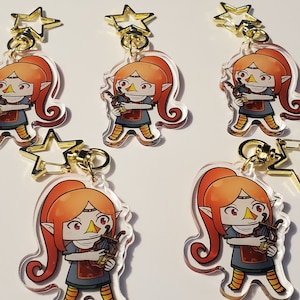 Puede incluir: Cuatro llaveros de acrílico transparente con un personaje de dibujos animados con cabello naranja, un vestido azul y un arpa. Cada llavero tiene un dije de estrella dorada.