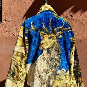 Puede incluir: Chaqueta de felpa de estilo vintage con un vibrante fondo azul y una detallada ilustración de un ciervo. Las mangas y el cuello presentan una mezcla de patrones florales amarillos y verdes, creando un diseño único y artístico.