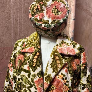 May include: A vintage floral velvet coat and matching hat. The coat features a cream background with large, detailed floral patterns in shades of pink, green, and gold. The hat mirrors the coat's design, creating a coordinated ensemble.