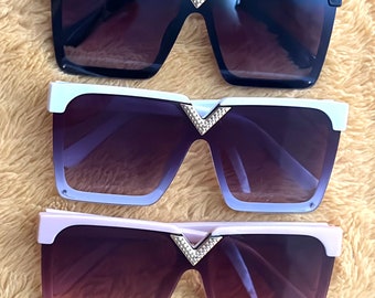 May include: Three pairs of square-shaped sunglasses with a V-shaped rhinestone detail on the bridge. The sunglasses are in black, white, and pink.