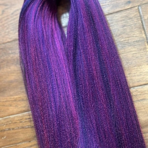 May include: A bundle of synthetic braiding hair in shades of purple, magenta, and black. The hair is tightly packed and appears ready for use in braids or other hairstyles. The colours blend together to create a vibrant and eye-catching effect.