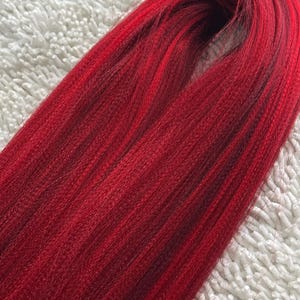 May include: A bundle of long, straight, red synthetic hair extensions. The hair is smooth and shiny, with a slight ombre effect from a darker red at the roots to a brighter red at the ends.