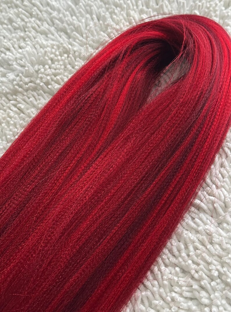 May include: A bundle of long, straight, red synthetic hair extensions. The hair is smooth and shiny, with a slight ombre effect from a darker red at the roots to a brighter red at the ends.