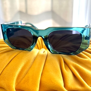 May include: Teal-colored sunglasses with dark lenses. The sunglasses have a thick, angular frame and are resting on a yellow, textured surface. The lenses are dark, providing contrast to the teal frame.