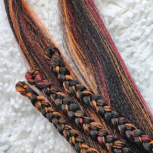 Op de afbeelding: Drie strengen zwart, oranje en rood haarverlenging zijn samen gevlochten. Het haar is lang en recht, en de vlechten zijn netjes gedaan.