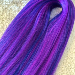 May include: A bundle of synthetic hair extensions in shades of purple and blue. The hair is straight and has a slight sheen.