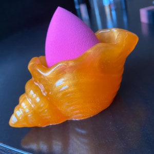 Può includere: Un porta spugne per trucco a forma di conchiglia dorata con una spugna per trucco rosa all'interno.