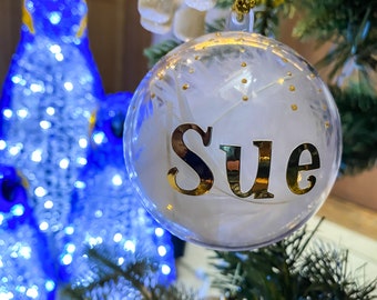 May include: A clear glass Christmas bauble with white feathers and gold glitter inside. The word "Sue" is written in gold letters on the bauble.
