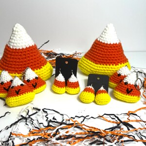 May include: A set of crocheted candy corn earrings, keychains, and ornaments. The candy corn is white, orange, and yellow. The earrings and keychains have smiling faces.
