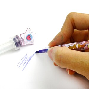 以下が含まれることがあります： 透明なプラスチック製のバレルと上部にハートのデザインが施された、紫とピンクの星型のペン。ペンは白い表面に描画するために使用されています。