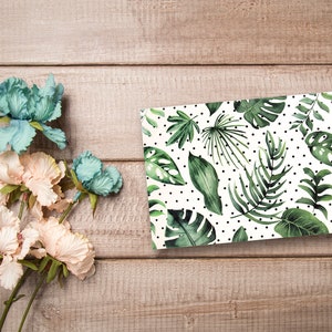 May include: A white card with a green tropical leaf pattern and black polka dots. The card is on a light brown wooden surface with a bouquet of light pink and blue flowers.
