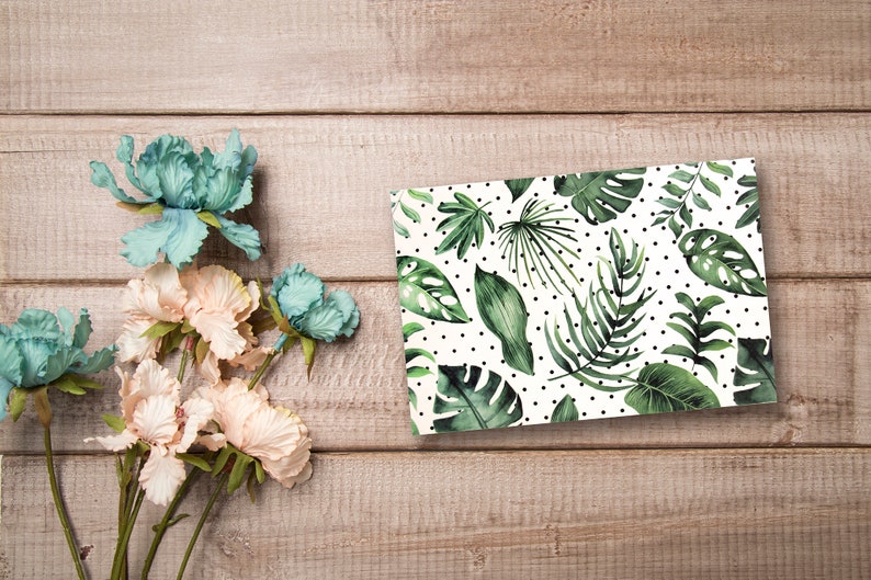 May include: A white card with a green tropical leaf pattern and black polka dots. The card is on a light brown wooden surface with a bouquet of light pink and blue flowers.