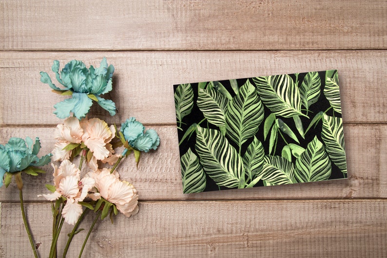 May include: A rectangular card with a tropical leaf pattern. The leaves are green and white on a black background.