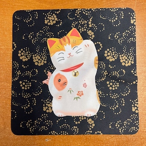 May include: A white and orange Maneki-Neko, or lucky cat, with a gold bell around its neck. The cat is sitting on a black background with a gold dot pattern.